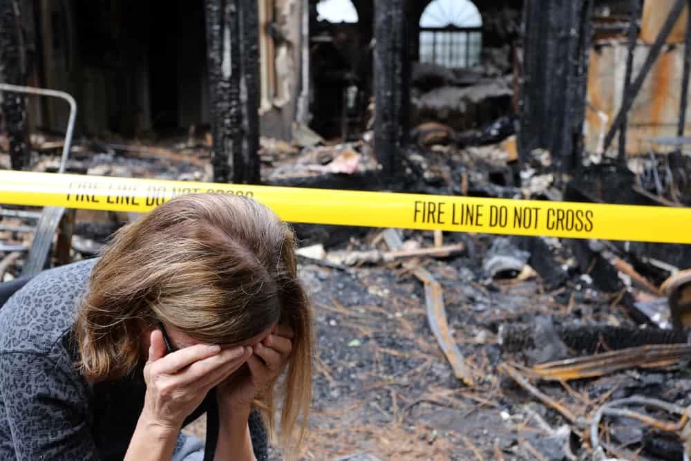 Woman sitting on the ground outside a fire-damaged building, looking distressed.