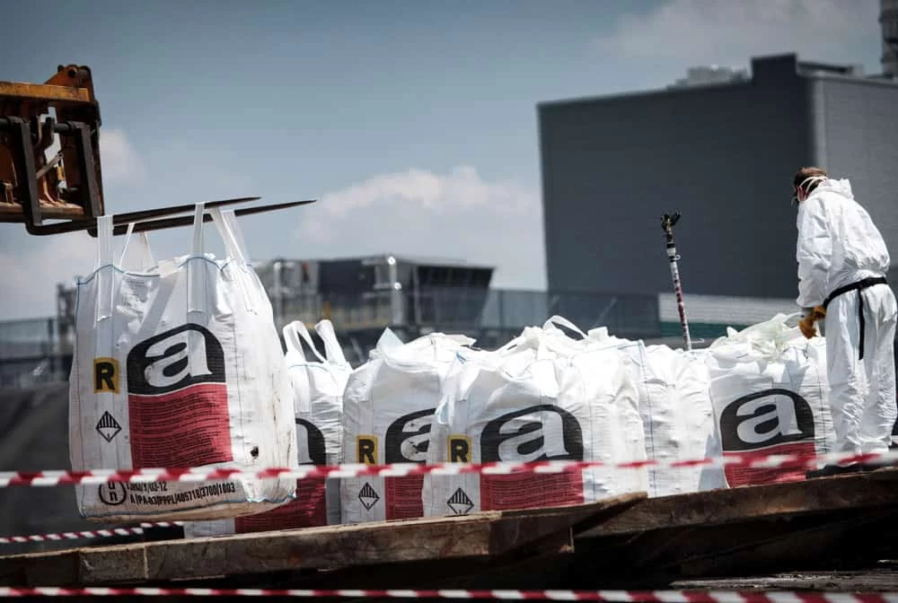 Worker wearing protective gear loading bags of chemicals in an industrial setting.