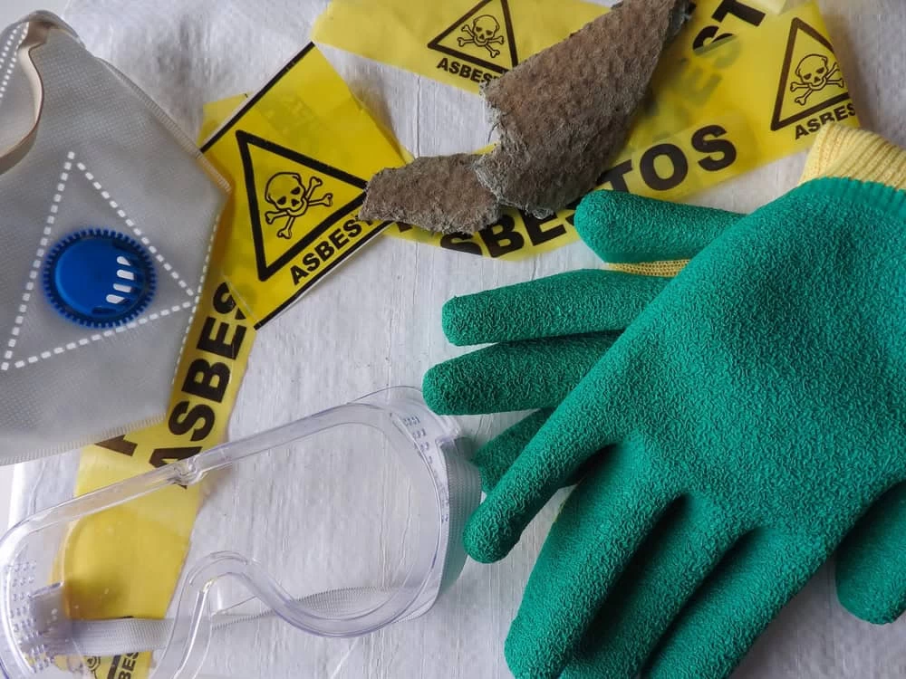 Protective gloves, goggles, and safety sign laid out for asbestos removal near roof tile samples.