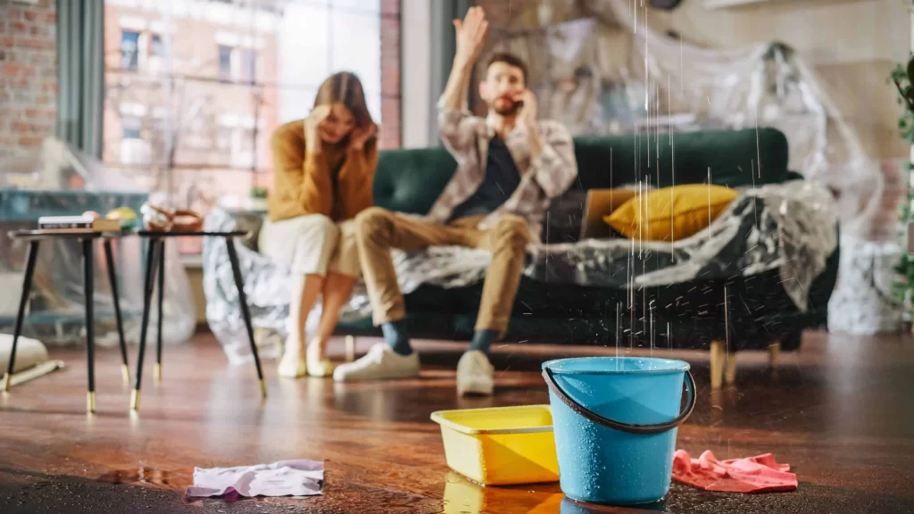 Frustrated couple making a phone call while dealing with a roof leak and burst pipe at home.